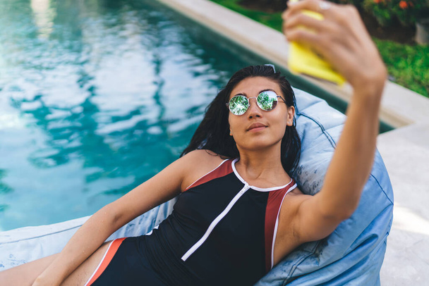 From above of tanned woman in sunglasses lying on beanbag lounge near swimming pool and taking selfie while resting on summer day - Foto, Bild