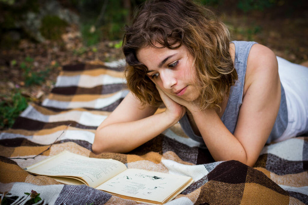 Beautiful teenager girl reading a book lying on blanket on green grass at the woods. - Zdjęcie, obraz