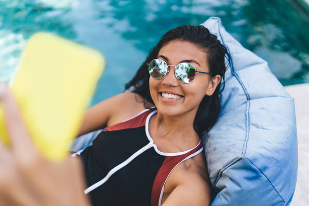 From above of smiling female in swimsuit relaxing on beanbag lounge and taking selfie while having rest near swimming pool - Foto, Imagem