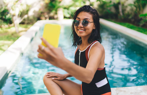 Attractive female with toothy smile in swimwear sitting on poolside with smartphone and taking selfie while having rest on sunny day - Fotografie, Obrázek