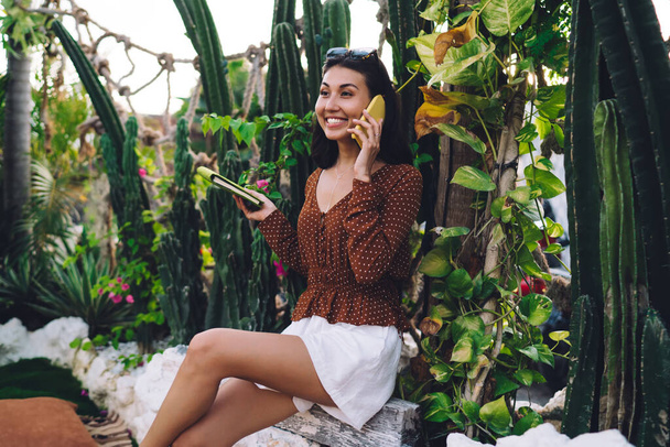 Cheerful young ethnic woman in casual outfit sitting on bench in exotic garden and having phone conversation against green tropical plants - Фото, изображение