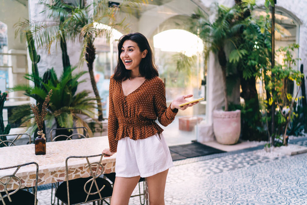Happy young Asian female with dark hair in stylish summer outfit standing near table with hand on chair and holding smartphone - Photo, Image