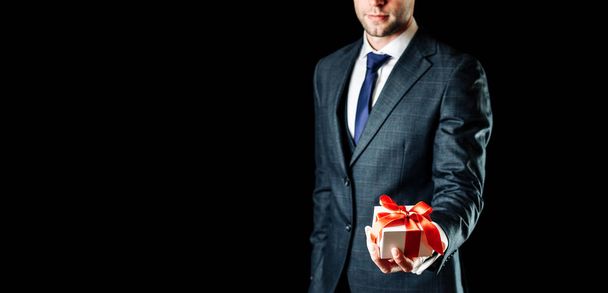 Ajándék van a kezében. Boldog fiatal üzletember kezében meglepetés ajándékdoboz jelen piros szalaggal elszigetelt fekete háttérrel. Születésnap, évfordulós ajándékok, ajándék kártya koncepció másolási hellyel - Fotó, kép