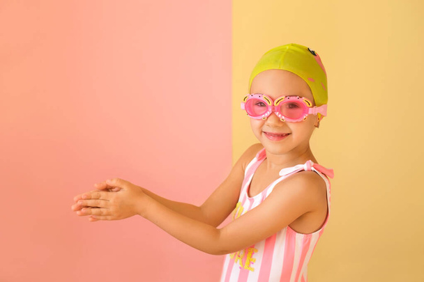 Een kinderzwemmer in een zwembril staat met zijn handen gevouwen in een houding om te zwemmen. Een meisje in een muts en gestreepte zwembroek om in het zwembad te zwemmen - Foto, afbeelding