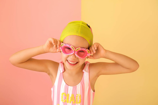 Un enfant en maillot de bain et lunettes et un bonnet de bain sourit doucement en regardant la caméra. Fille nageuse Rose et jaune fond studio. - Photo, image