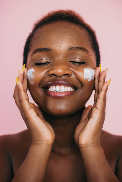 Closeup portrait of an african american woman applying hydration cream on her smiling face, isolated over pink background. Clean skin. Body care. - Foto, Imagen