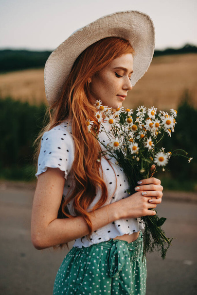 Redhead woman with a large bouquet of wildflowers walking along rural road - Фото, изображение