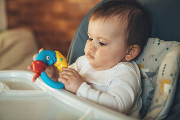 Caucasian newborn baby playing with colorful educational rattle toy while sitting on baby table waiting the meal. - Фото, изображение