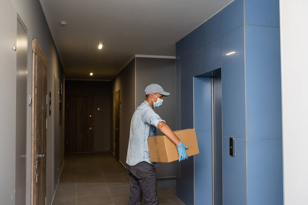 Kotiinkuljetus laatikko mies yllään käsineet ja suojaava naamio toimittaa paketteja ovella - Valokuva, kuva