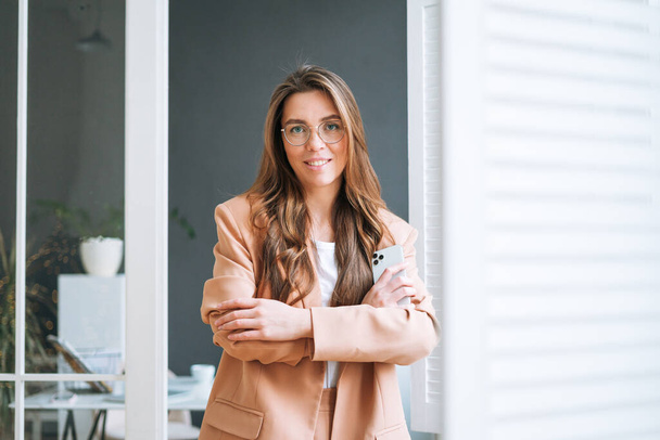 Young smiling brunette business woman in glasses with long hair in stylish beige suit using smartphone in the modern office - Foto, afbeelding