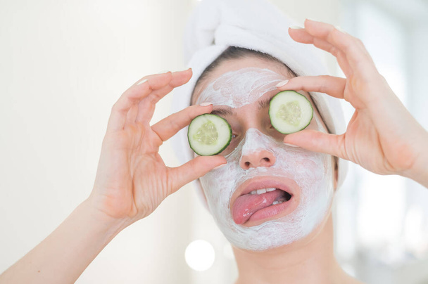 Cheerful woman with a towel on her hair and in a clay face mask fooling around with cucumbers in her hands. Taking care of beauty at home - 写真・画像