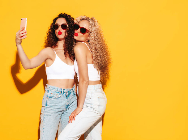 Two beautiful sexy smiling hipster women in red summer swimwear bathing suits. Trendy models with afro curls hairstyle having fun in studio. Hot female isolated on yellow in sunglasses. Taking selfie - Foto, Imagem
