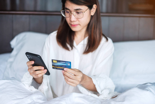 自宅で朝にベッドで注文しながらオンラインショッピングに携帯電話やクレジットカードを使う女性。テクノロジーeコマースデジタルバンキングオンライン決済アパート生活の概念 - 写真・画像