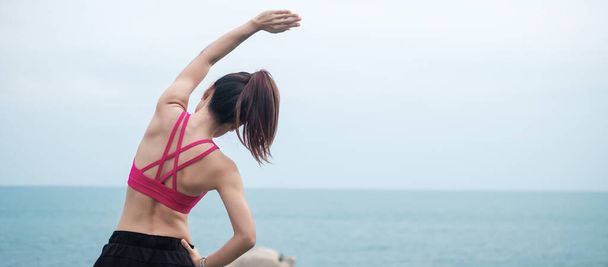 Jonge fitnessvrouw in sportkleding stretching lichaam tegen uitzicht op de oceaan, gezonde vrouw oefening in de ochtend. Workout, wellness en work life balance concepten - Foto, afbeelding