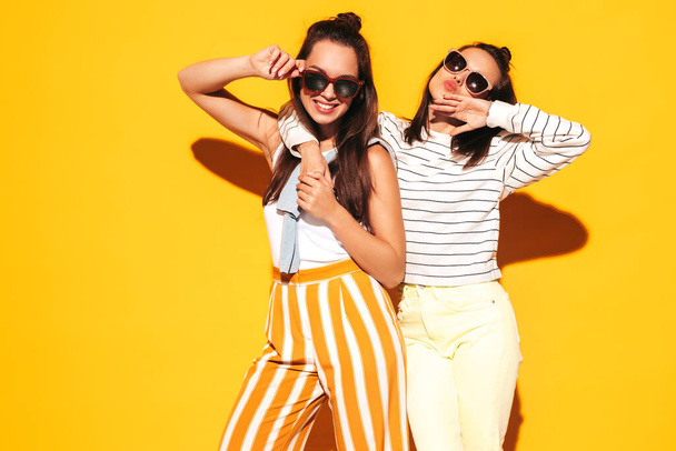 Two young beautiful smiling blond hipster female in trendy summer clothes. Sexy carefree women posing near yellow wall in studio. Positive models having fun. Cheerful and happy. In sunglasses - Foto, immagini