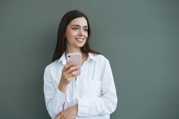 Portrait of beautiful smiling woman student with dark long hair in white shirt using mobile phone in the hand on grey background isolated, generation Z - Фото, изображение