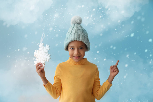 Cute little African-American boy in winter clothes and with snowflake showing something on color background with falling snow - Fotografie, Obrázek
