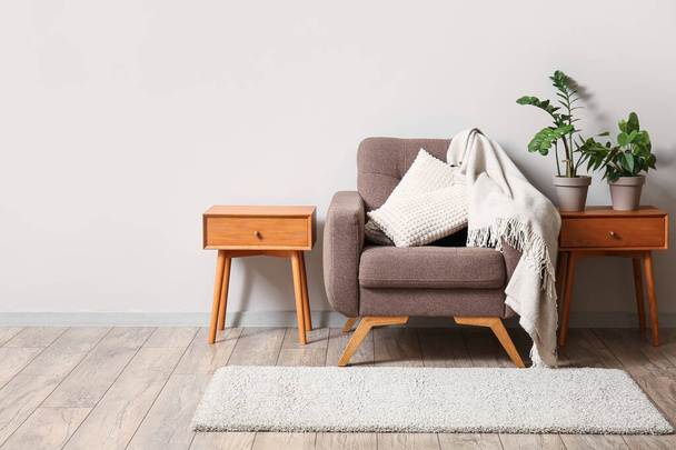 Zachte fauteuil en houten tafels met kamerplanten bij lichte wand - Foto, afbeelding