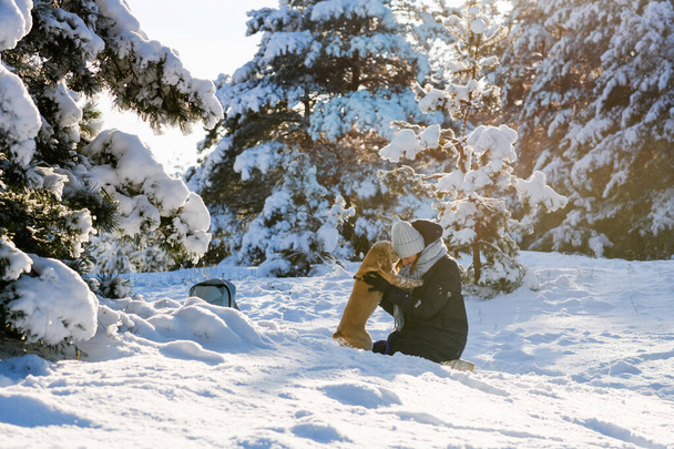 La niña se sienta de rodillas en una deriva de nieve y el perro se para en sus dos patas traseras. La chica abraza a su mascota. Mujer joven con Cocker Spaniel americano en el bosque de invierno. - Foto, imagen