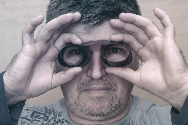A man looking through binoculars made of vintage film. Two hands hold a twisted photographic film in front of his eyes. A close-up. Selective focus. - Foto, Bild