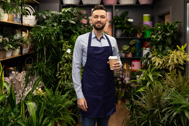 florist entrepreneur in his shop surrounded by flowers and potted plants stands with a cup of coffee and looks with a smile at the camera. - Photo, Image