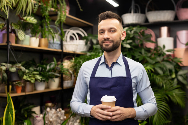 a florist in a blue apron with a cup of coffee in his hands stands in a flower and bouquet shop and looks at the camera with a smile. - Foto, Bild