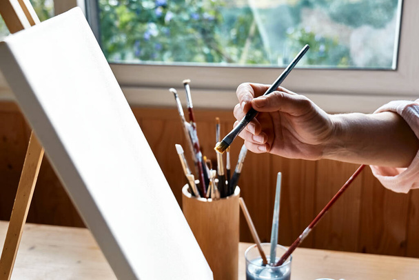Vrouwelijke hand met een penseel in de buurt van doek op ezel bij het raam in de studio. Kunstschilderkunst hobby. Creatieve vrije tijd. Zelfexpressie concept. - Foto, afbeelding