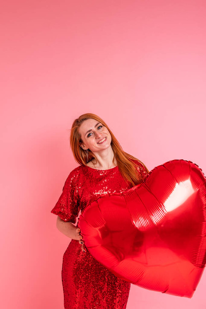 Beautiful redhead girl with red heart baloon posing. Happy Valentine's Day concept. Studio photo of beautiful ginger girl dancing on pink background. - 写真・画像