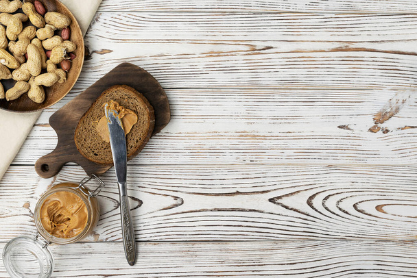 A piece of bread spread with peanut butter on a dark brown wooden board. Peanuts in a wooden saucer on a light table. Peanut paste in a glass jar. Top view, space for text - Fotoğraf, Görsel