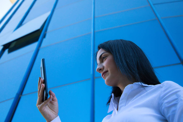 young, beautiful woman is consulting her mobile phone at the entrance door of the building where she works. The photo is taken from below and the building can be seen. Concept businesswoman. - Foto, Bild
