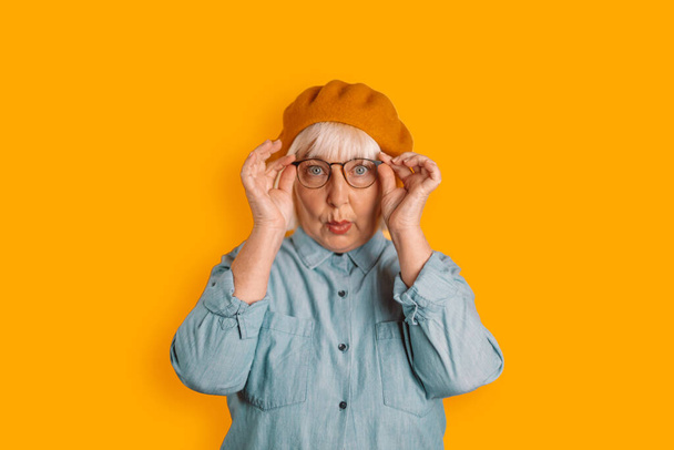 Photo of blonde hair 50s woman astonished face touch glasses look camera isolated over orange color background - Photo, Image
