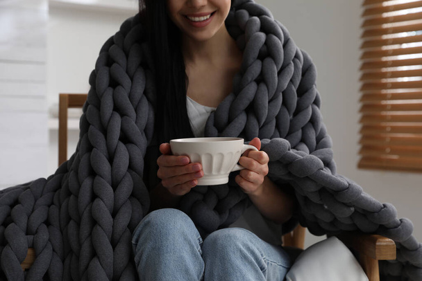 Woman with chunky knit blanket and cup in armchair at home, closeup - 写真・画像