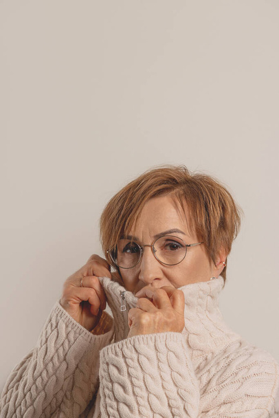 Beautiful middle-aged baby boomer woman in glasses wrapped in beige sweater on neutral background. - Fotografie, Obrázek