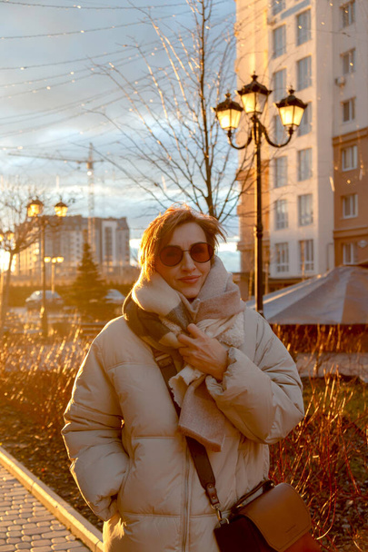 Beautiful baby boomer woman of middle age in sunglasses and stylish casual clothes on the street. Against background of sunset. - Zdjęcie, obraz