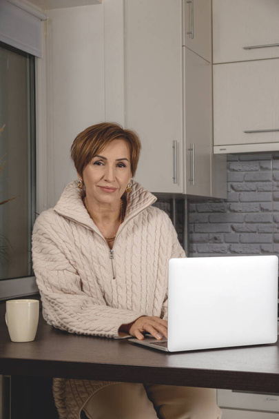 Beautiful middle-aged baby boomer woman working in laptop. Background of light gray interior. - Foto, Bild