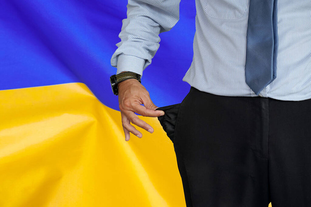 Gros plan de la main de l'homme en costume tournant la poche de son pantalon à l'envers et le montrant vide sur le fond du drapeau ukrainien. Crise, chômage, concept de faillite en Ukraine - Photo, image