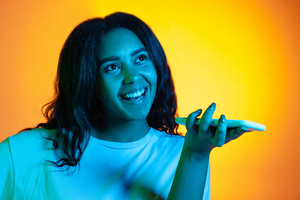 Close-up cheerful young pretty dark skinned girl wearing white t-shirt using phone isolated on yellow background in neon light, filter. - Foto, Imagem