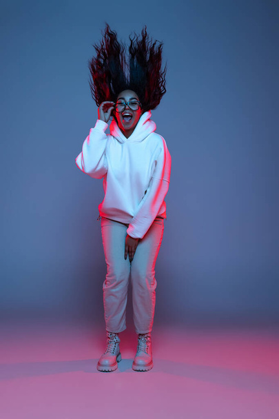 Full-length portrait of young happy dark skinned girl in hoodie jumping, dancing isolated on gradient blue-pink background in neon. - Foto, Imagem