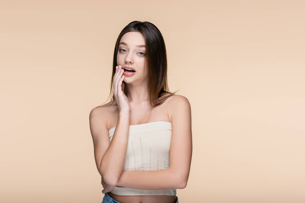 young woman in crop top with bare shoulders and open mouth looking at camera isolated on beige - Foto, Imagen