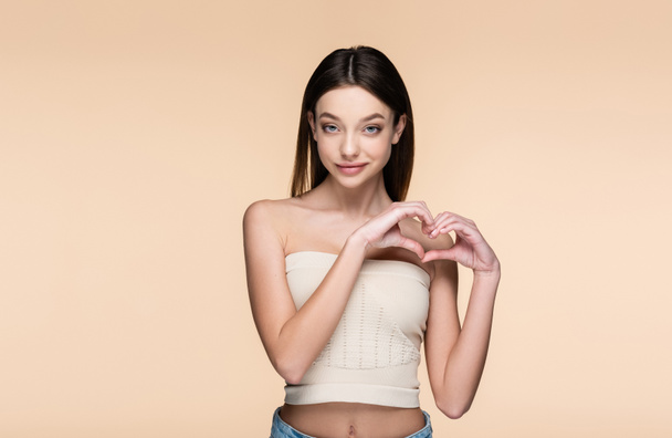smiling and pretty woman in crop top showing heart with hands isolated on beige  - Photo, Image