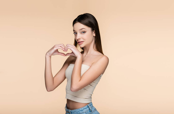brunette and pretty woman in crop top showing heart with hands isolated on beige  - Foto, imagen