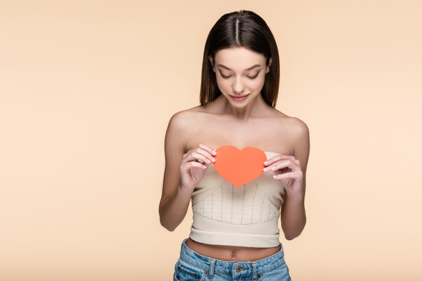 brunette woman in crop top with bare shoulders looking at red paper heart isolated on beige  - Foto, Imagem