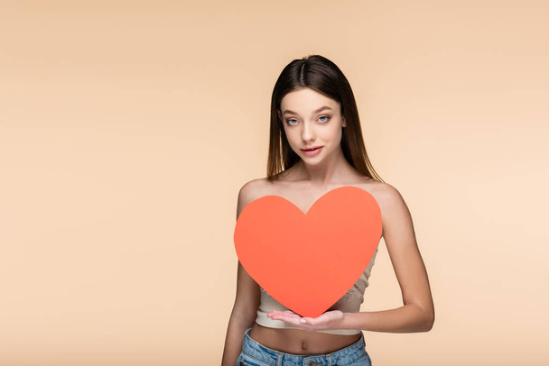 young brunette woman with bare shoulders holding red paper heart isolated on beige  - Fotó, kép