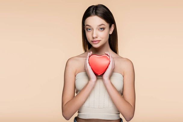 brunette young woman holding red heart-shaped metallic box isolated on beige  - 写真・画像