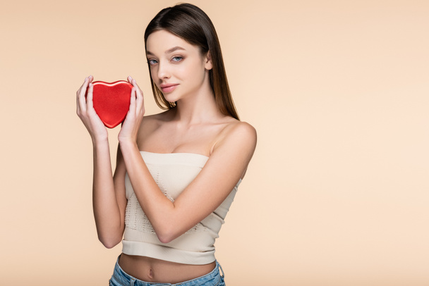 brunette young woman holding heart-shaped metallic box isolated on beige  - 写真・画像