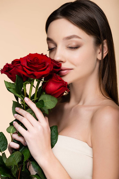 brunette young woman with closed eyes smelling red roses isolated on beige  - Foto, Imagen