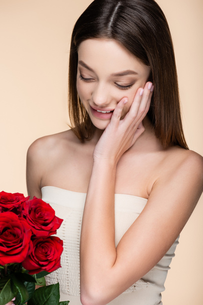 cheerful woman with bare shoulders looking at bouquet of red roses isolated on beige  - Foto, immagini