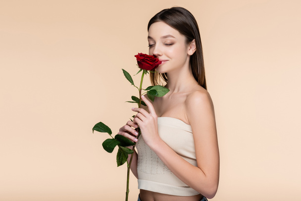 young woman with closed eyes smelling red rose isolated on beige  - Fotó, kép