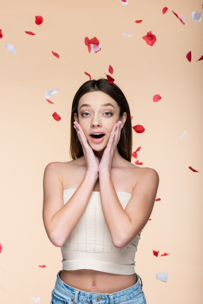 excited young woman in crop top near falling rose petals on beige  - Photo, Image
