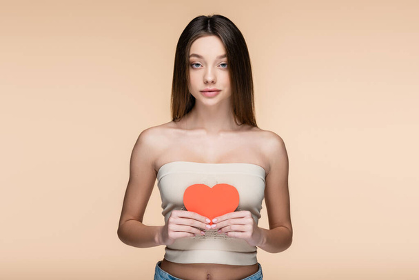 sensual woman in crop top with bare shoulders holding red paper cut heart isolated on beige - Foto, imagen